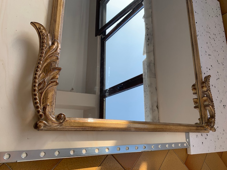 Op de afbeelding: Een sierlijke gouden spiegel met een decoratief, gerold frame. De spiegel reflecteert een raam met uitzicht op de lucht.