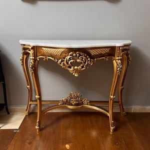 May include: A gold-painted console table with a white marble top and intricate carvings. The table has a curved top and ornate legs.