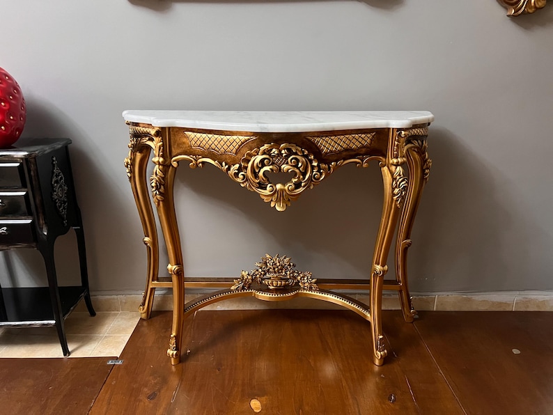 May include: A gold-painted console table with a white marble top and intricate carvings. The table has a curved top and ornate legs.
