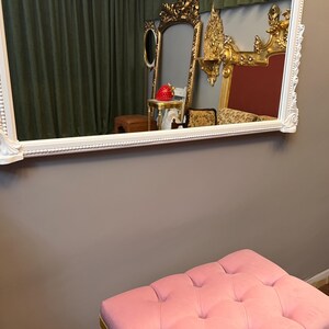 Op de afbeelding: Een witte, sierlijke spiegel met een roze, gestoffeerde poef er voor. De spiegel reflecteert een kamer met gouden meubels en een rode muur.