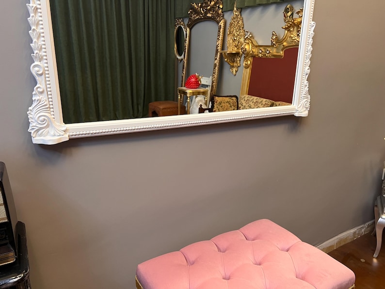 Op de afbeelding: Een witte, sierlijke spiegel met een roze, gestoffeerde poef er voor. De spiegel reflecteert een kamer met gouden meubels en een rode muur.