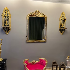 Peut inclure: Un miroir doré avec des détails ornés est accroché à un mur gris, encadré par deux étagères murales noires et dorées. Un canapé chaise longue en velours rose se trouve devant le miroir, et une statue dorée d'une femme se tient sur une table d'appoint noire.