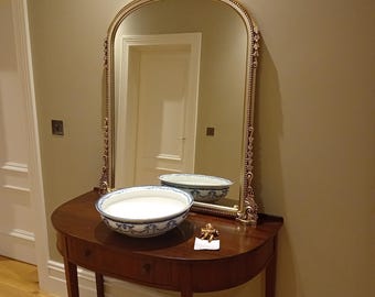 May include: A semi-circular wooden console table with a drawer, topped with a white ceramic basin featuring blue floral patterns. Behind the table stands an arched mirror with a gold-toned frame decorated with ornate leaf and scroll carvings.