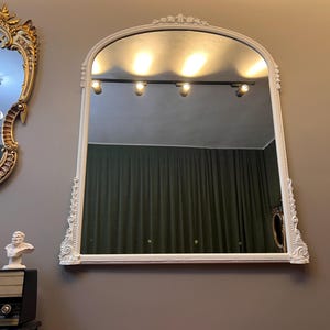 Op de afbeelding: Een witte, sierlijke, gebogen spiegel met een gefacetteerde rand en een decoratief frame. De spiegel reflecteert een kamer met een groen gordijn en een railsysteem voor verlichting.
