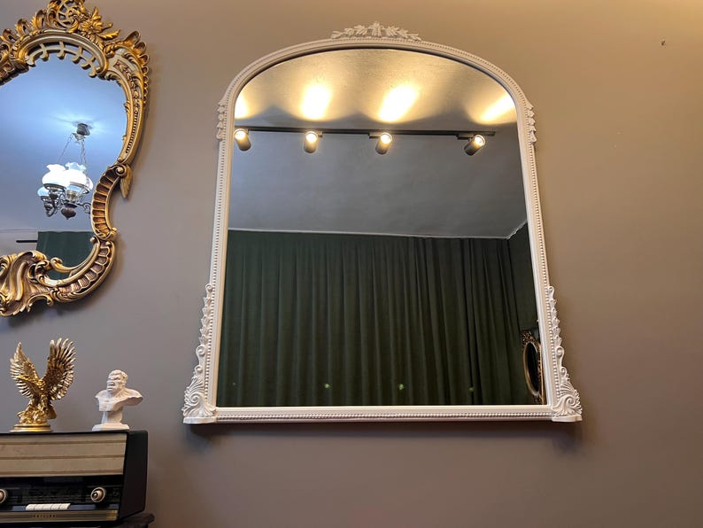 Op de afbeelding: Een witte, sierlijke, gebogen spiegel met een gefacetteerde rand en een decoratief frame. De spiegel reflecteert een kamer met een groen gordijn en een railsysteem voor verlichting.
