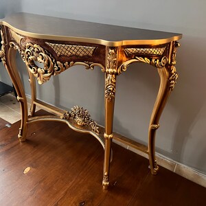 May include: A gold-painted console table with ornate carvings and a dark brown top. The table has a curved front and a lower shelf.