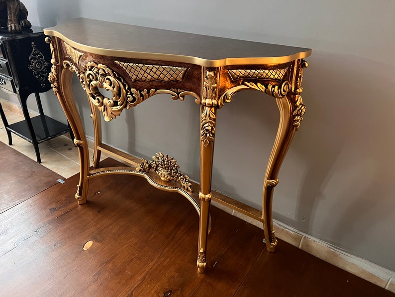 May include: A gold-painted console table with ornate carvings and a dark brown top. The table has a curved front and a lower shelf.