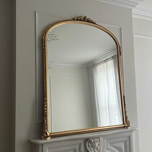 Op de afbeelding: Een grote, sierlijke, gouden spiegel met een boogvorm hangt boven een witte marmeren open haard. De spiegel heeft een decoratief ontwerp rond de lijst en een parelrand.