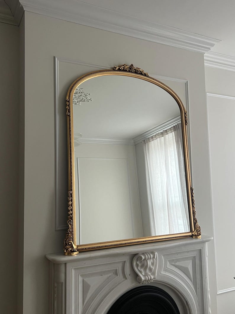 Op de afbeelding: Een grote, sierlijke, gouden spiegel met een boogvorm hangt boven een witte marmeren open haard. De spiegel heeft een decoratief ontwerp rond de lijst en een parelrand.