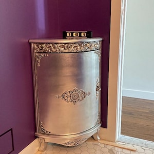 May include: A silver corner cabinet with ornate details and a single door. The cabinet is set against a purple wall.