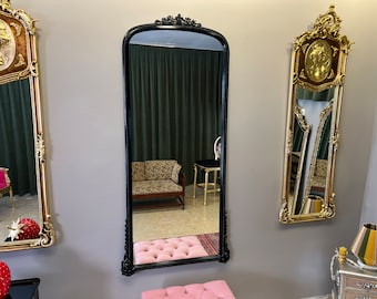 May include: A black ornate mirror with a gold-colored frame and a reflection of a room with a couch and a chair. The mirror is hanging on a gray wall.