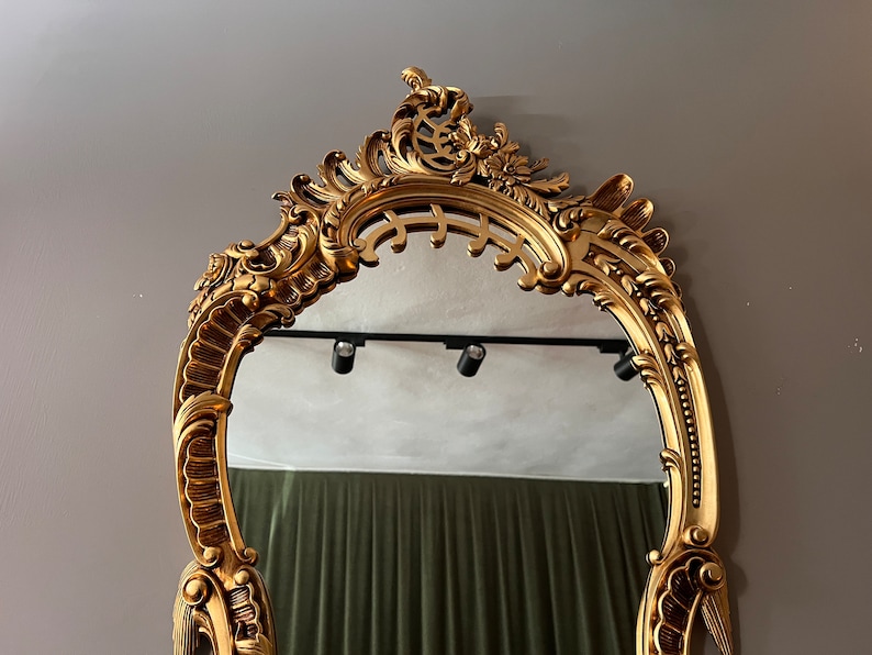 May include: A gold ornate mirror with a decorative, intricate frame. The mirror is oval shaped and has a beveled edge.