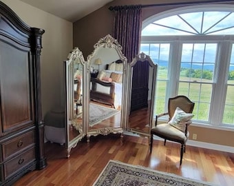 May include: A large, dark wood armoire with ornate details stands in a bedroom with a three-panel mirrored screen, a chair, and a window with a view of a green field.