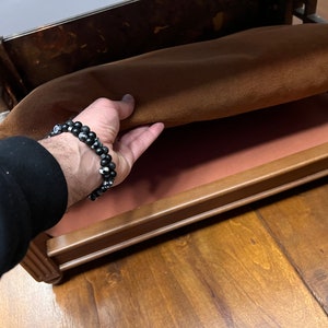 May include: A wooden bed frame with a brown fabric mattress. The mattress is being lifted to reveal a pink fabric lining underneath.