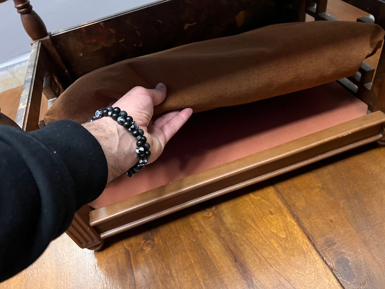 May include: A wooden bed frame with a brown fabric mattress. The mattress is being lifted to reveal a pink fabric lining underneath.