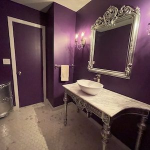 Könnte beinhalten: Ein weißes Porzellanwaschbecken mit einem silbernen Wasserhahn steht auf einer Marmor-Arbeitsplatte mit verzierten silbernen Beinen. Das Waschbecken befindet sich in einem Badezimmer mit violetten Wänden und einem silbernen Spiegel mit verzierten Details.