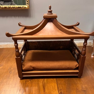 May include: A wooden dog bed with a canopy top. The bed is brown and has a brown cushion. The bed is on a wooden floor.