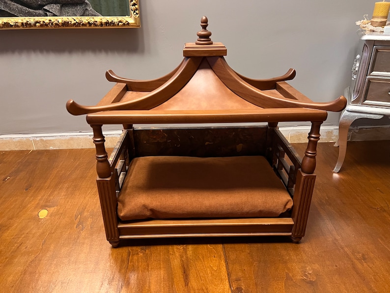 May include: A wooden dog bed with a canopy top. The bed is brown and has a brown cushion. The bed is on a wooden floor.