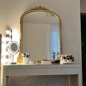Op de afbeelding: Een witte kaptafel met een gouden spiegel met sierlijke details. De spiegel reflecteert een kamer met witte muren en een raam met witte jaloezie&euml;n. De kaptafel heeft een glazen blad en een witte lade. Er staan ​​verschillende flessen en potten op de tafel, evenals een make-upkwast houder.