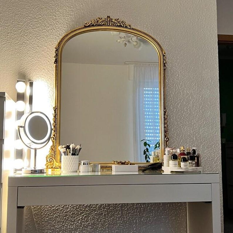 Op de afbeelding: Een witte kaptafel met een gouden spiegel met sierlijke details. De spiegel reflecteert een kamer met witte muren en een raam met witte jaloezie&euml;n. De kaptafel heeft een glazen blad en een witte lade. Er staan ​​verschillende flessen en potten op de tafel, evenals een make-upkwast houder.