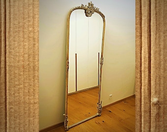 May include: A tall, ornate gold-framed mirror with a decorative top. The mirror is standing in a room with wooden floors and white walls.