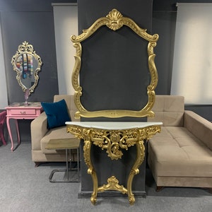May include: Ornate gold-colored console table with a white marble top, paired with a matching gold-framed mirror. The table features intricate carvings and curved legs. A sofa and other decorative furniture are in the background.