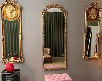 May include: Three ornate gold-framed mirrors mounted on a grey wall. The two outer mirrors feature decorative gold relief portraits at the top, while the central mirror has an arched top. Each frame displays intricate scrollwork and floral detailing.