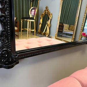 Op de afbeelding: Een zwarte sierlijke spiegel met een decoratief frame. De spiegel reflecteert een kamer met meubels en een raam.
