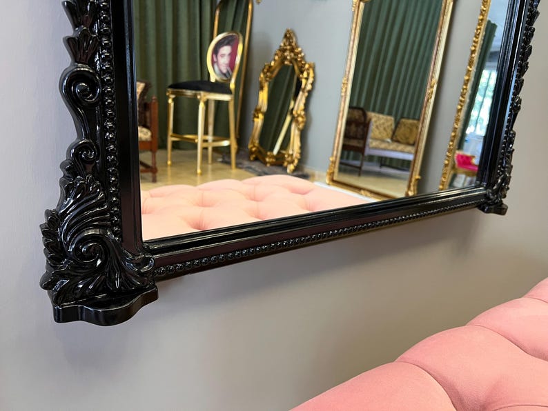 Op de afbeelding: Een zwarte sierlijke spiegel met een decoratief frame. De spiegel reflecteert een kamer met meubels en een raam.