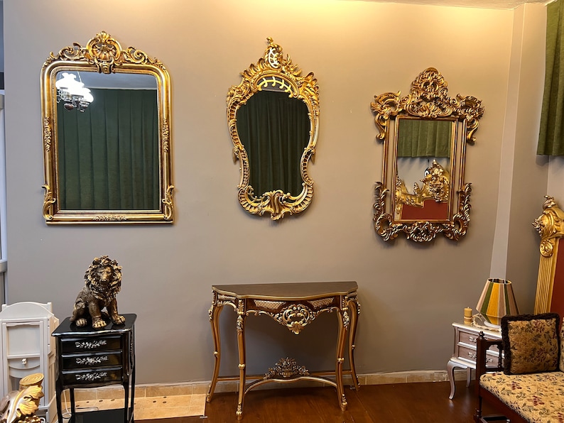 May include: Three ornate gold-framed mirrors with intricate carvings hang on a wall above a dark brown console table with gold accents. A gold lion statue sits on a black side table with three drawers.