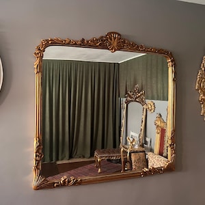 May include: A large, ornate gold-framed mirror with intricate carvings hangs on a gray wall. A smaller, round white mirror and a gold wall sconce are also visible. The mirror reflects a room with a chair and curtains.