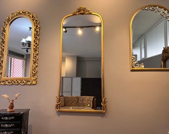 May include: Three ornate gold-framed mirrors with intricate carvings. The largest mirror is standing upright and reflects the room's interior. The two smaller mirrors are hanging on the wall.