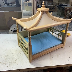 May include: A wooden dog bed with a light blue cushion and a pagoda-style roof. The bed is made of light wood with gold accents and has a decorative lattice design on the sides.