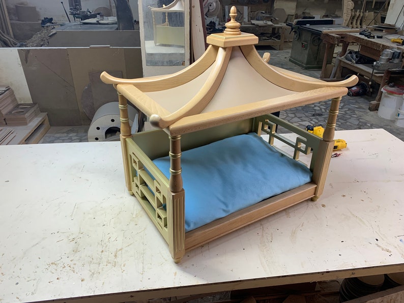 May include: A wooden dog bed with a light blue cushion and a pagoda-style roof. The bed is made of light wood with gold accents and has a decorative lattice design on the sides.
