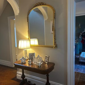 Op de afbeelding: Een gouden, gebogen spiegel met sierlijke details hangt aan een lichtbeige muur boven een donkerhouten consoletafel. Op de tafel staat een witte lamp met een cr&egrave;mekleurige kap, een decoratieve ster en een paar ingelijste foto's.