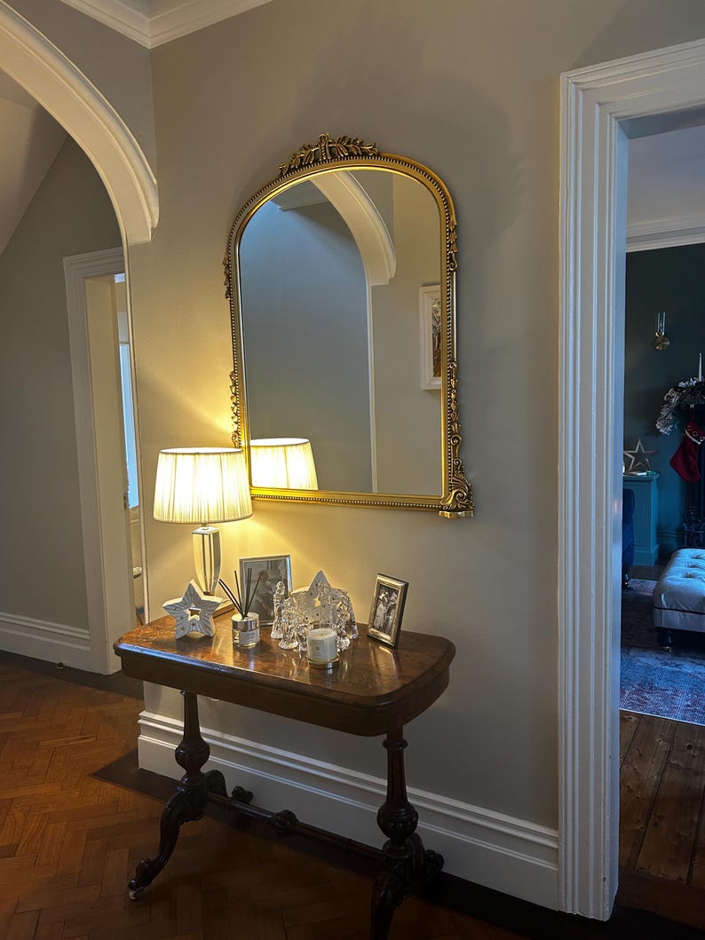 Op de afbeelding: Een gouden, gebogen spiegel met sierlijke details hangt aan een lichtbeige muur boven een donkerhouten consoletafel. Op de tafel staat een witte lamp met een cr&egrave;mekleurige kap, een decoratieve ster en een paar ingelijste foto's.