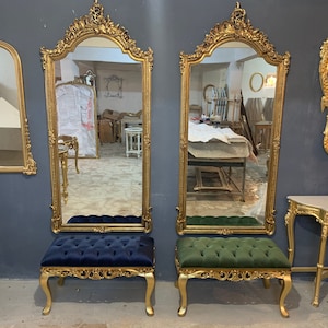 May include: Ornate gold-framed mirrors with matching upholstered benches. The mirrors have a detailed, scrolling design and are paired with tufted benches in navy blue and green. The furniture is set against a dark gray wall.