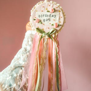 May include: A pink and cream-coloured 'birthday girl' rosette with the text "birthday girl". The rosette is decorated with small pink roses, gold stars, and green and pink ribbons. The rosette is approximately 13 cm in diameter.