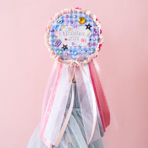 May include: A circular badge featuring blue and white gingham fabric with the embroidered text birthday girl. The badge is decorated with colorful sequins, rhinestones, and stars, finished with a ruffled lace edge and hanging pink and white ribbons.