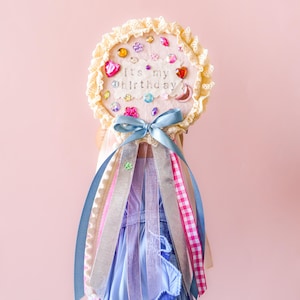 May include: A decorative birthday wand with a cream-colored, lace-edged circle. The circle is adorned with colorful gems and the text "it's my birthday". Blue, pink, and silver ribbons cascade from the wand.
