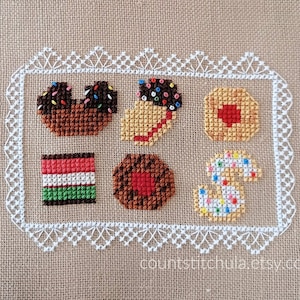 May include: A cross-stitch design of a plate of cookies, including a chocolate cookie with sprinkles, a gingerbread cookie, a sugar cookie with sprinkles, a heart-shaped cookie, a cookie with red icing, and a gingerbread cookie shaped like a number 3. The design is framed with a white lace border.