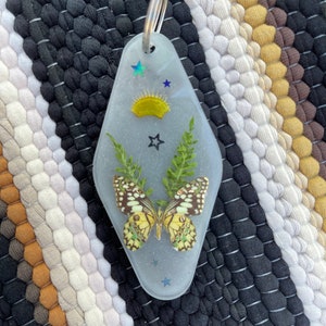 May include: A clear resin keychain shaped like a motel key with a pressed butterfly, green leaves, and glitter. The keychain also features a yellow sun and blue stars.