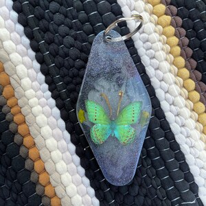 May include: A green butterfly with yellow accents is encased in a clear, diamond-shaped keychain with a silver ring.