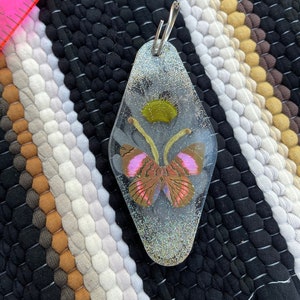 May include: A clear resin keychain with a glittery finish, featuring a pressed butterfly with pink and brown wings. The butterfly is surrounded by a green flower.