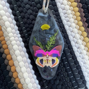May include: A black and silver motel key chain with a pink and yellow butterfly and green foliage pressed inside. The key chain is shaped like a motel key.