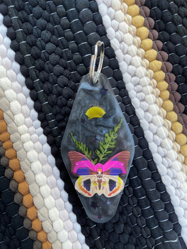 Peut inclure: Un porte-cl&eacute;s de motel noir et argent avec un papillon rose et jaune et un feuillage vert press&eacute; &agrave; l'int&eacute;rieur. Le porte-cl&eacute;s est en forme de cl&eacute; de motel.