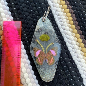 May include: A silver key chain with a clear resin charm featuring a pink and brown butterfly with glitter inside. The charm is shaped like a motel key.