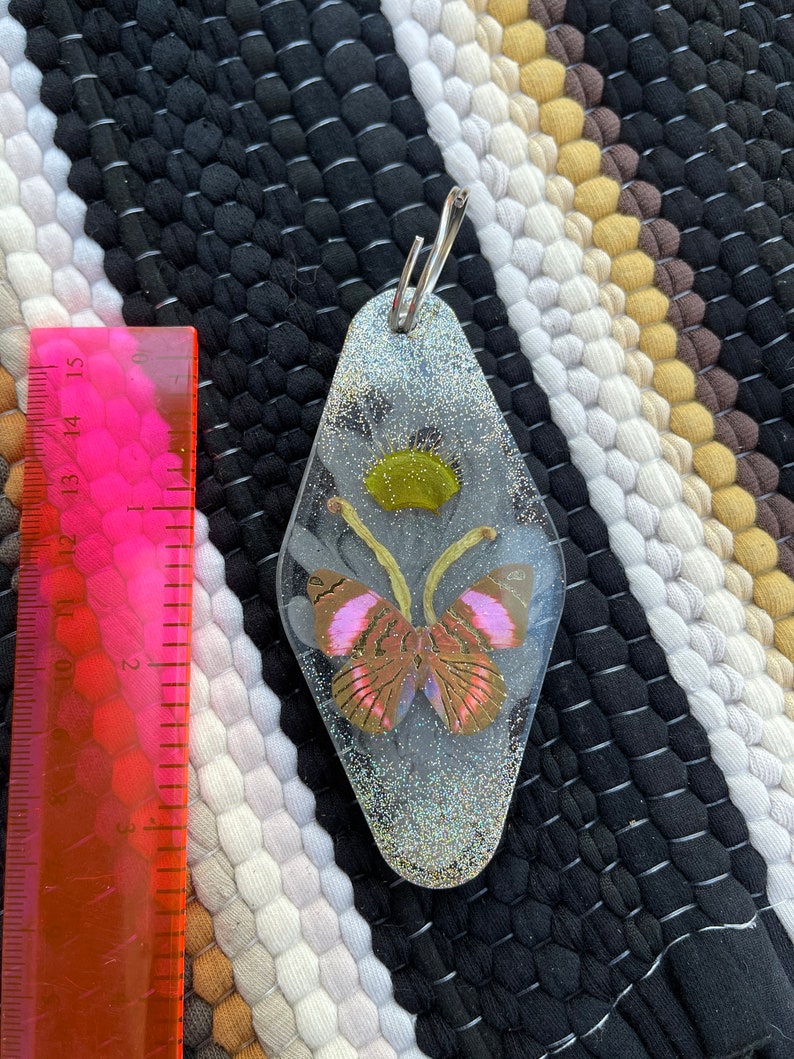 May include: A silver key chain with a clear resin charm featuring a pink and brown butterfly with glitter inside. The charm is shaped like a motel key.