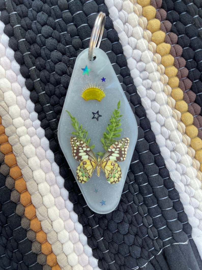 Op de afbeelding: Een transparante sleutelhanger van hars in de vorm van een motel-sleutel met een geperste vlinder, groene bladeren en glitter. De sleutelhanger heeft ook een gele zon en blauwe sterren.