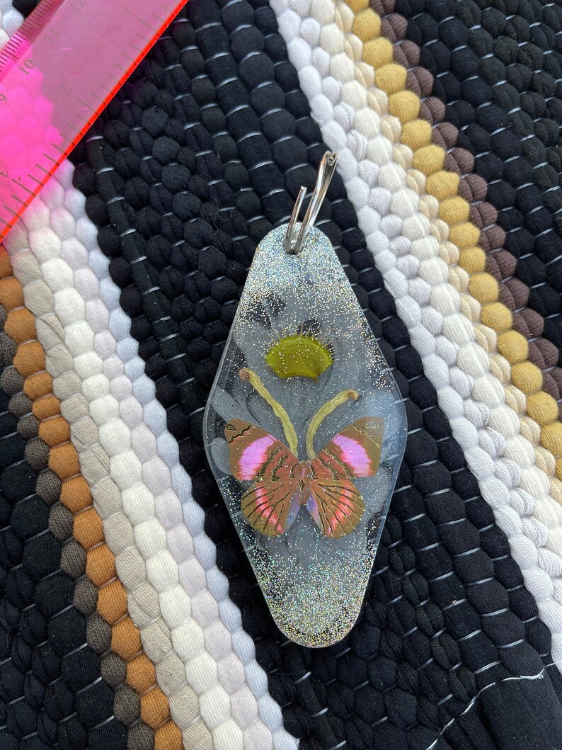 May include: A clear resin keychain with a glittery finish, featuring a pressed butterfly with pink and brown wings. The butterfly is surrounded by a green flower.